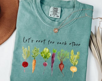 May include: A teal Comfort Colors t-shirt with the phrase "Let's root for each other" and watercolor illustrations of various root vegetables. A silver necklace with a pendant and a brown hat are also visible.