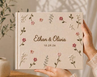 May include: A rectangular beige photo album with embroidered floral motifs in pink, white, and green. The cover features the text "Ethan & Olivia" in dark brown script, followed by the date "10.29.26" in a simple, clean font.