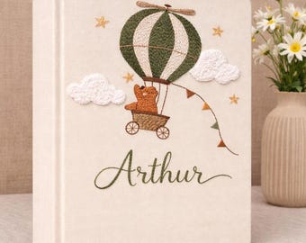 May include: A cream-colored fabric-covered book featuring an embroidered design of a brown bear in a hot air balloon with green and white stripes. The scene includes white clouds, small orange stars, and the name Arthur stitched in green script.