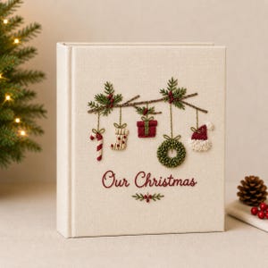 Op de afbeelding: Een cr&egrave;mekleurig, met stof bekleed fotoalbum met geborduurde kerstversieringen zoals een zuurstok, sok, cadeau, krans en kerstmuts die aan een tak hangen. De tekst "Our Christmas" is in het rood onder de versieringen geborduurd.