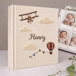 Może przedstawiać: Kremowy album fotograficzny z haftowanymi detalami. Okładka przedstawia samolot, chmury, balon na ogrzane powietrze i imię "Henry" w ozdobnym kroju pisma. Album jest otwarty, ukazując zdjęcia dziecka.