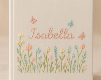 May include: A square photo album with a cream-colored fabric cover featuring embroidered pastel wildflowers in shades of pink, yellow, and blue. The name Isabella is stitched in pink cursive script in the center, accented by three small embroidered butterflies.