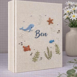 May include: A beige fabric-covered book featuring embroidered sea creatures and the name "Ben" in blue script. The design includes a blue whale, starfish, crab, fish, and seaweed. Bubbles and small dots complete the underwater scene.