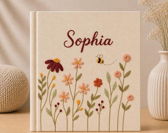 May include: A square, cream-colored fabric-covered book featuring the embroidered name Sophia in dark red script. The cover is decorated with embroidered wildflowers in shades of pink, red, and yellow, along with a small bee and a dotted flight path.