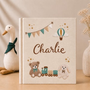 Pode incluir: Uma capa de livro retangular de tecido cor de creme com o nome Charlie bordado em letra cursiva castanha. O design inclui uma grinalda de bandeirolas azul-petróleo e bege, um balão de ar quente, um urso de peluche e um comboio.
