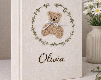 May include: A cream-colored fabric-covered photo album featuring an embroidered design of a brown teddy bear with a light blue ribbon around its neck, centered within a green leaf wreath. The name Olivia is embroidered in dark brown script below.