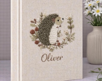 May include: A beige fabric-covered photo album featuring an embroidered design of a hedgehog surrounded by small flowers and leaves. The name Oliver is embroidered in brown script below the hedgehog. The album has a vertical spine detail.