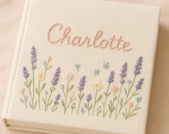 May include: A square, cream-colored fabric-covered photo album featuring the name Charlotte embroidered in pink script. The cover is decorated with embroidered wildflowers, including purple lavender sprigs, white daisies, and small pink and blue buds.