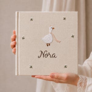 Op de afbeelding: Een vierkant, met beige stof bekleed fotoalbum met een geborduurde witte gans die een roze sjaal draagt. De naam Nora is in donkerbruin cursief onder de gans geborduurd, omringd door kleine geborduurde bloemtakjes in de hoeken.