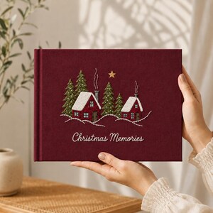 Op de afbeelding: Een donkerrood, met stof bekleed fotoalbum met een geborduurde wintersc&egrave;ne van twee rode huizen, groene dennenbomen, witte sneeuw en een gouden ster. De tekst "Christmas Memories" is in witte letters op de voorkant geborduurd.