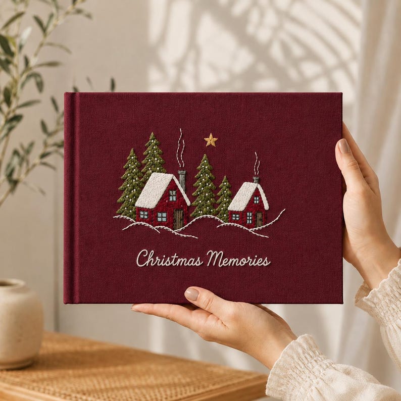 Op de afbeelding: Een donkerrood, met stof bekleed fotoalbum met een geborduurde wintersc&egrave;ne van twee rode huizen, groene dennenbomen, witte sneeuw en een gouden ster. De tekst "Christmas Memories" is in witte letters op de voorkant geborduurd.