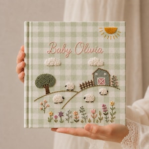 Op de afbeelding: Een vierkant fotoalbum met een lichtgroene en witte gingham stoffen kaft. De voorkant bevat geborduurde details waaronder een gele zon, witte wolken, een schuur, een boom, schapen en kleurrijke bloemen. De tekst Baby Olivia is in het roze geborduurd.