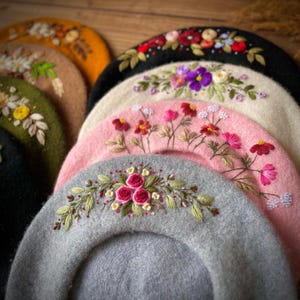 Könnte beinhalten: Eine Kollektion von Wollbaskenmützen in verschiedenen Farben wie Grau, Rosa, Creme, Schwarz, Olivgrün und Senfgelb. Jede Baskenmütze ist mit aufwendigen, handgestickten Blumenmotiven verziert, die mit farbigen Fäden Rosen und Wildblumen darstellen.