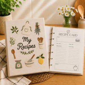 Könnte beinhalten: Ein cremefarbener Rezeptordner aus Stoff mit gestickten Küchenmotiven wie einer Schürze, einem Schneebesen, einer Zitrone und einer Waage. Der Text "My Recipes" ist in Schwarz gestickt. Der geöffnete Ordner zeigt eine bedruckte weiße Rezeptkarte mit Feldern für Titel, Zubereitungszeit, Kochzeit, Zutaten und Anweisungen.