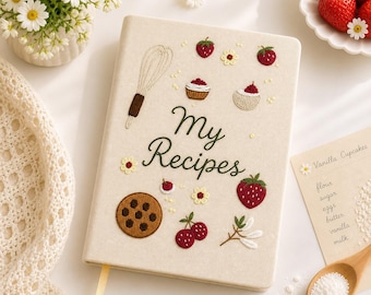 Puede incluir: Un cuaderno de recetas beige con ilustraciones bordadas de fresas, un batidor, una galleta y las palabras "My Recipes". El cuaderno tiene una cinta amarilla de marcador y está rodeado de margaritas y fresas.