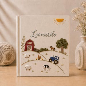 Pode incluir: Um livro revestido de tecido cor de creme com uma cena de quinta bordada, incluindo um celeiro vermelho, um trator azul, um cavalo, uma vaca, ovelhas e uma galinha. O nome Leonardo está bordado em letra cursiva cinzento-escura sobre a paisagem com um sol amarelo.