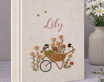 May include: A cream-colored fabric-covered photo album featuring embroidered details of a wheelbarrow filled with orange flowers, two small birds, and two bees. The name Lily is embroidered in pink script above the wheelbarrow design.