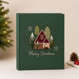 Op de afbeelding: Een donkergroen hardcover boek met een geborduurde wintersc&egrave;ne van een huis met een bordeauxrood dak, omringd door groene bomen en witte sneeuwvlokken. De tekst Merry Christmas is in wit cursief schrift onder het huis geborduurd.