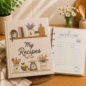 May include: A cream-coloured fabric recipe binder featuring embroidered kitchen motifs including shelves, a scale, and flowers. The cover displays the text "My Recipes" in dark brown script. An open page shows a printed "RECIPE CARD" template.