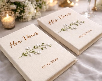 May include: Two cream-coloured vow books with embroidered floral details. The text "Her Vows" and "His Vows" are in brown script. Each book features a floral design and the date "03.12.2026" in brown. Wedding theme.