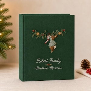 Op de afbeelding: Een donkergroen fotoalbum met een geborduurd ontwerp van een konijn in een trui dat een gestreepte kous aan een dennentak hangt. De kaft bevat de geborduurde tekst Robert Family en Christmas Memories in een wit cursief lettertype.