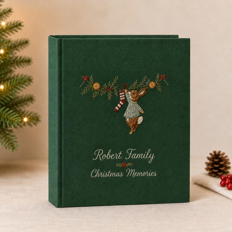 Op de afbeelding: Een donkergroen fotoalbum met een geborduurd ontwerp van een konijn in een trui dat een gestreepte kous aan een dennentak hangt. De kaft bevat de geborduurde tekst Robert Family en Christmas Memories in een wit cursief lettertype.