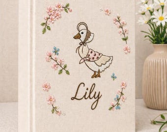 May include: A cream-colored book with an embroidered design featuring a duck wearing a bonnet and a pink scarf. The name "Lily" is embroidered below the duck. Pink and blue floral embroidery surrounds the duck.
