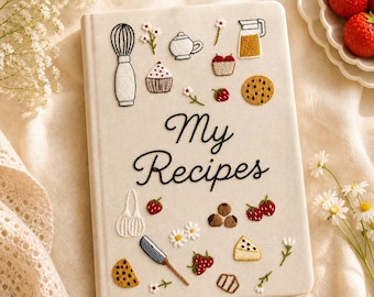 Puede incluir: Libro de recetas color crema con diseños bordados con temática de cocina, incluyendo un batidor, tetera, magdalena y fresas. Las palabras "My Recipes" están escritas en cursiva. Una cinta amarilla como marcapáginas es visible.
