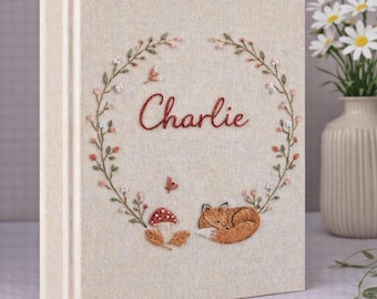 May include: A cream-colored fabric-covered photo album featuring an embroidered wreath of small flowers and leaves. The center displays the name Charlie in red script, with a small embroidered red mushroom and a sleeping orange fox below.
