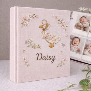 Op de afbeelding: Een lichtroze fotoalbum met een geborduurd ganzenontwerp en de naam "Daisy". De omslag heeft bloemenborduurwerk rond de gans. Het album is open en toont foto's van een baby.