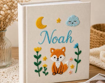 May include: A cream-colored fabric photo album featuring embroidered designs of a yellow crescent moon, a small star, a blue smiling cloud, and a fox. The name Noah is embroidered in blue script in the center, surrounded by green floral stems.