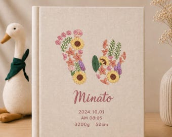 May include: A beige fabric-covered memory book featuring embroidered floral patterns in the shape of a footprint and a handprint. The text reads Minato, 2024.10.01, AM 08:05, 3200g, 52cm. The embroidery uses yellow, pink, purple, and green thread.