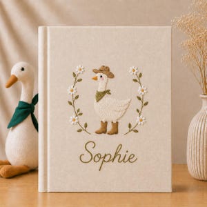 May include: A cream-coloured book featuring an embroidered goose wearing a cowboy hat, boots, and a neckerchief, encircled by a floral wreath. The name "Sophie" is embroidered below. A stuffed goose toy with a green scarf is to the left.