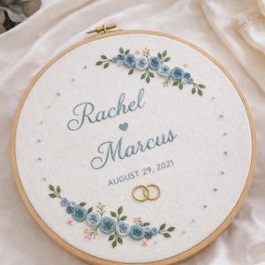 Könnte beinhalten: Gestickter Stickrahmen mit den Namen "Rachel" und "Marcus" und dem Datum "29. August 2021". Das Design zeigt blaue Blumenakzente und zwei goldene Eheringe. Die Stickerei befindet sich in einem Holzrahmen.