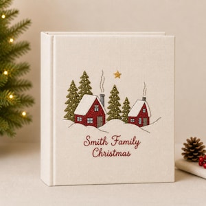 Op de afbeelding: Een cr&egrave;mekleurig stoffen fotoalbum met een geborduurde wintersc&egrave;ne met twee rode huizen, groene dennenbomen en een gouden ster. De tekst "Smith Family Christmas" is in rood cursief geborduurd onder het besneeuwde landschapsontwerp.
