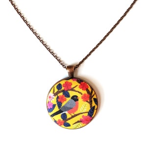 May include: A yellow, round pendant necklace with a copper chain. The pendant features a colorful floral design with a gray and red bird perched on a branch.