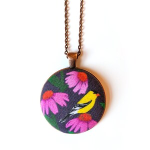 Pode incluir: Um colar com um pingente redondo e uma corrente de cobre. O pingente apresenta um design de tecido de um pássaro amarelo com asas pretas e uma flor rosa sobre um fundo roxo escuro.