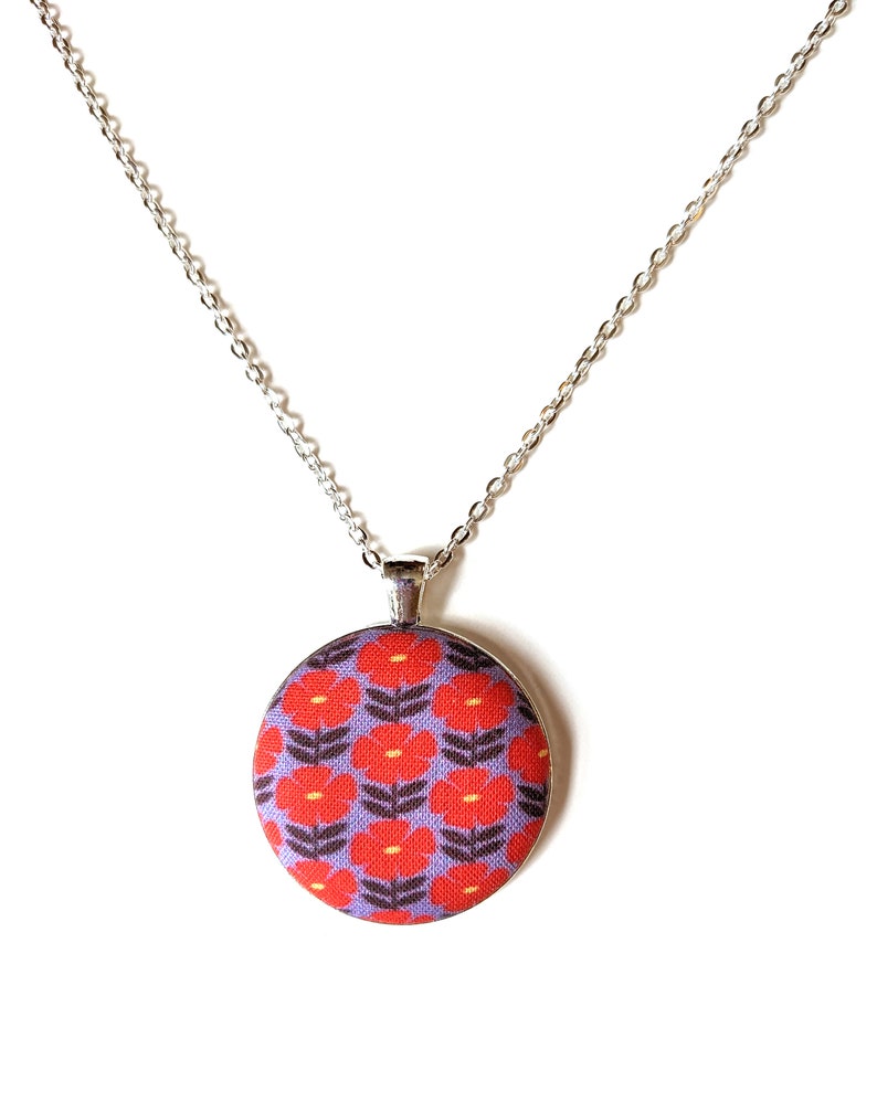 The fabric pendant necklace is hanging from a silver colored chain. The pendant has a light purple background with a pattern of red flowers.