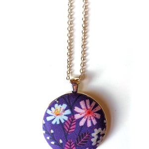 The circle shaped pendant hangs from a silver colored chain. The pendant is purple with pink, black, and white flowers and leaves.