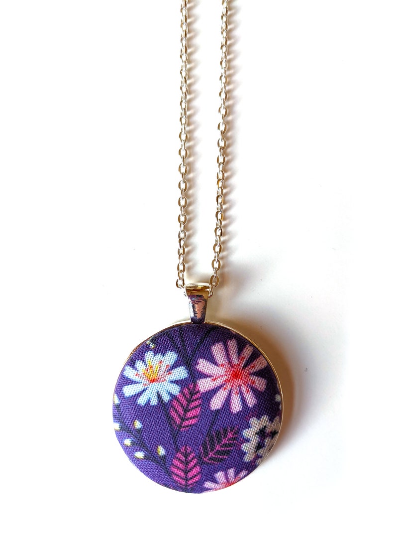 The circle shaped pendant hangs from a silver colored chain. The pendant is purple with pink, black, and white flowers and leaves.