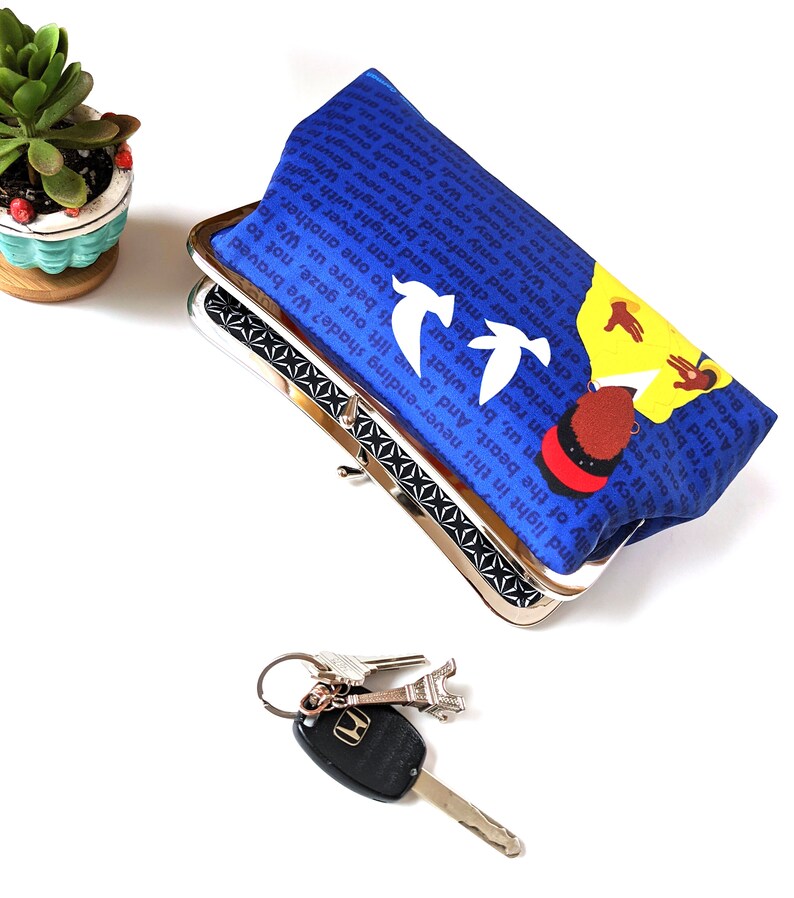 The Amanda Gorman clutch purse is shown from above, slightly opened. There is a set of keys on front and a plant off to the side. The inside of the clutch is black with a white design.