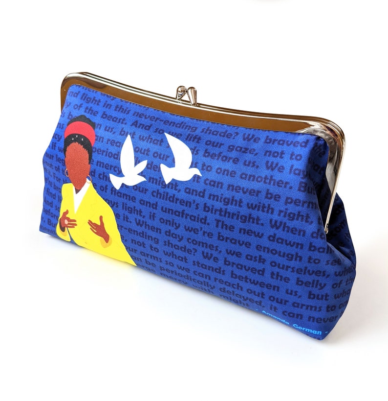 A clutch purse on a white background. The clutch is blue with dark blue text in the background. Amanda Gorman is shown wearing a yellow coat and a red band in her hair. There are two white flying birds next to her.