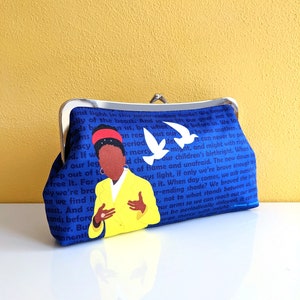 The clutch purse is on a white table with a yellow background behind it. The purse is primarily blue with a picture of Amanda Gorman and white birds. The clutch has a silver kiss lock clasp at the top.