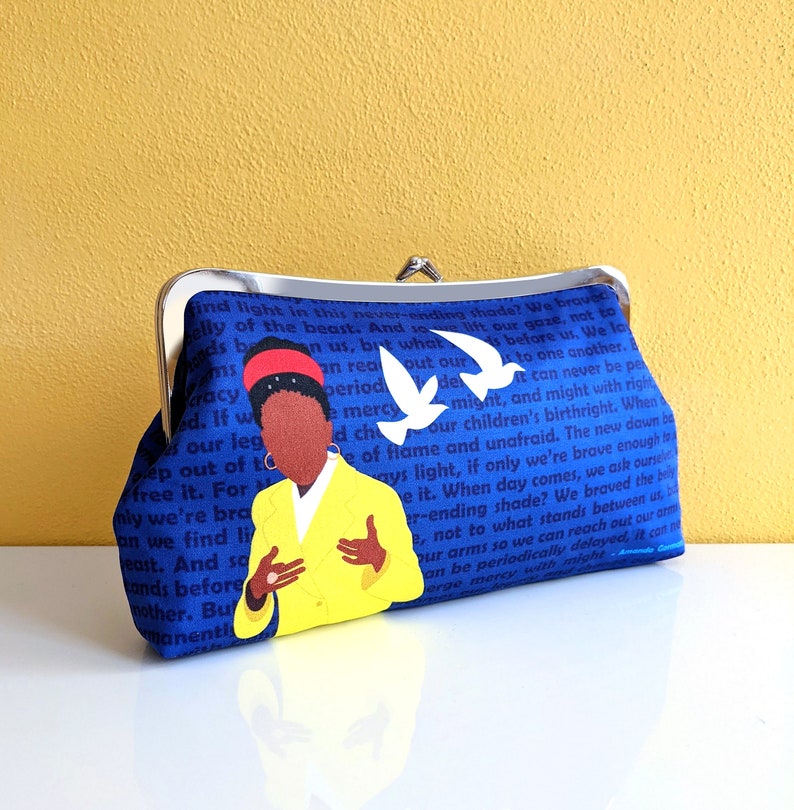 The clutch purse is on a white table with a yellow background behind it. The purse is primarily blue with a picture of Amanda Gorman and white birds. The clutch has a silver kiss lock clasp at the top.