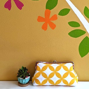 The yellow and white clutch is shown next to a plant against a yellow floral wall.