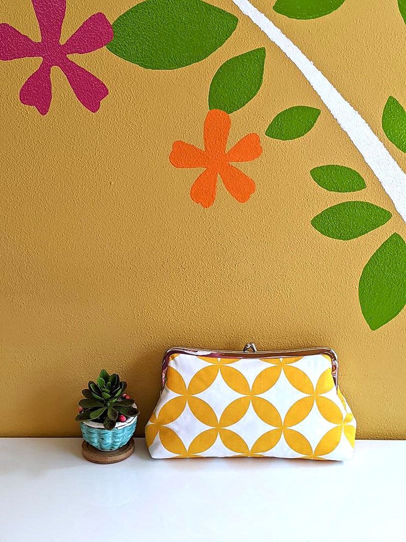 The yellow and white clutch is shown next to a plant against a yellow floral wall.