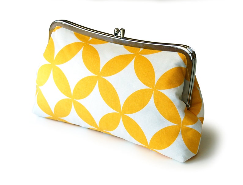 The yellow and white geometric clutch sits on a white background, slightly tilted to the side. The clutch has a silver colored clasp at the top.