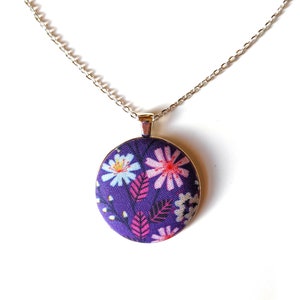 The circle shaped pendant hangs from a silver colored chain. The pendant is purple with pink, black, and white flowers and leaves.