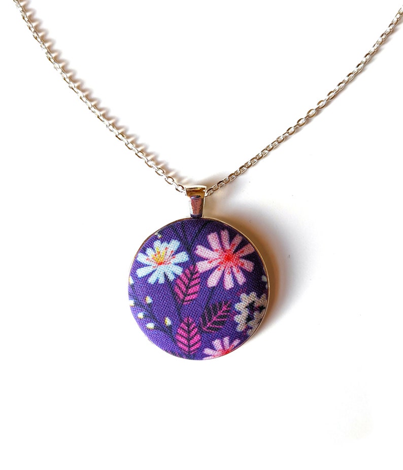 The circle shaped pendant hangs from a silver colored chain. The pendant is purple with pink, black, and white flowers and leaves.