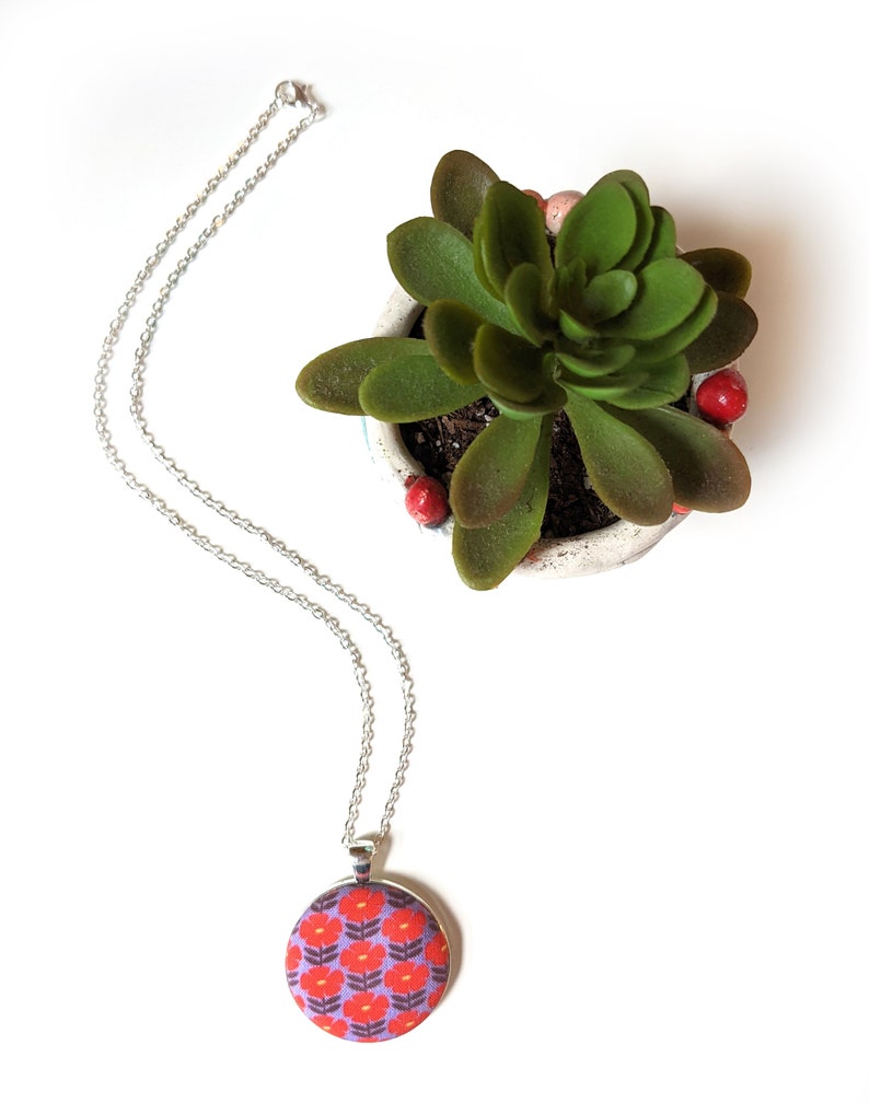 The floral fabric necklace is shown from above on a white background, next to a green plant.